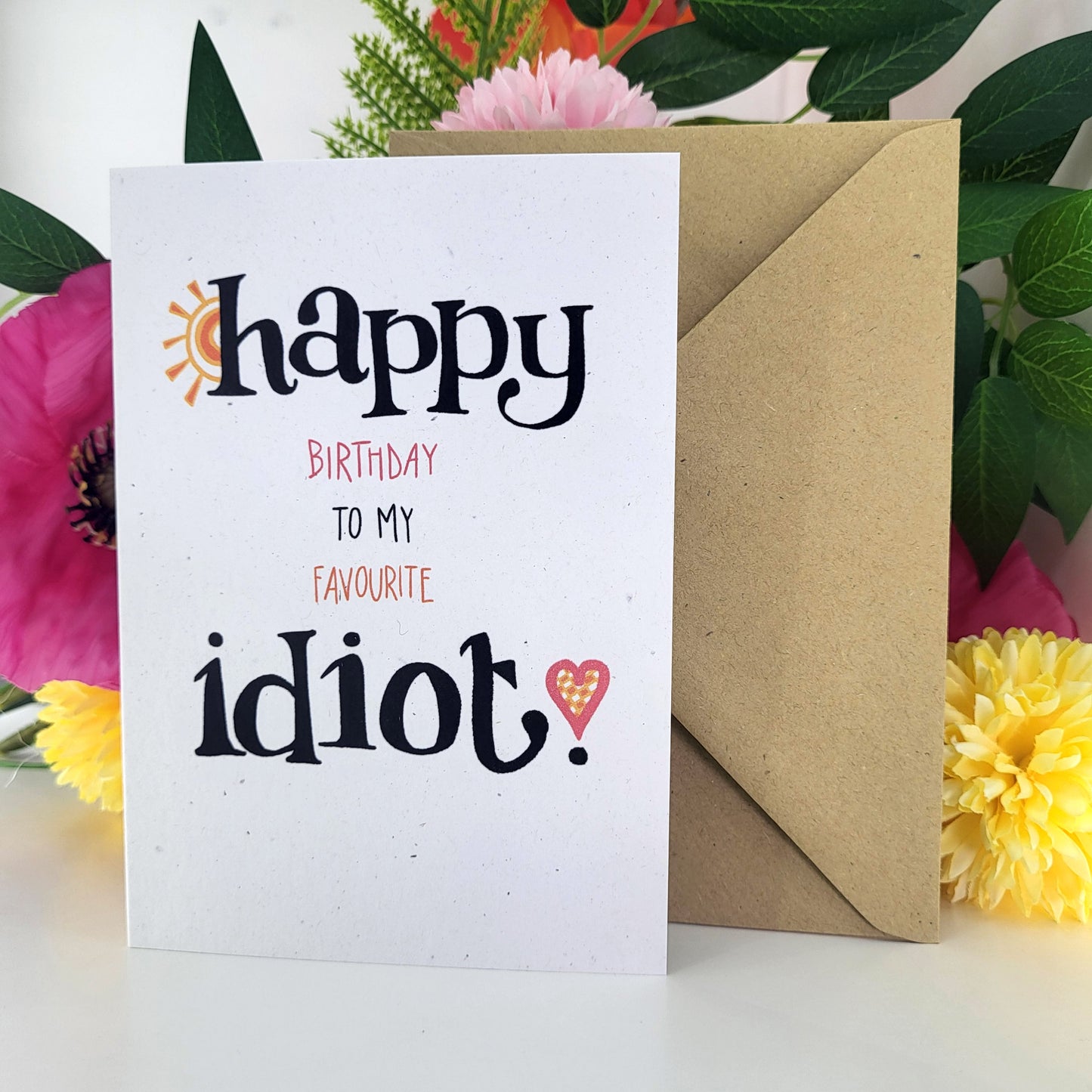 A white greeting card with the text 'happy birthday to my favourite idiot.' in black and pink lettering, shown with a brown kraft envelope behind it and colorful flowers around the edges.