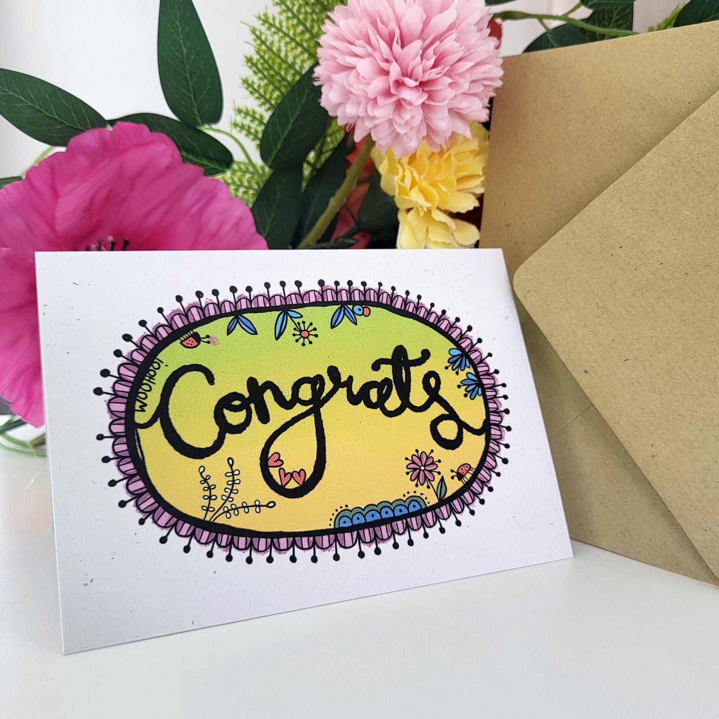 A white greeting card with a colorful oval design reading 'Congrats' in black swirling script, styled with pink and yellow flowers and brown envelope on a white surface.