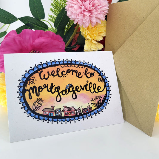 A greeting card with a hand-drawn oval design reading 'Welcome to Mortageville' in black script, surrounded by blue floral border details and a painterly sunset and small illustrated houses, displayed with a kraft envelope and flowers in the background.