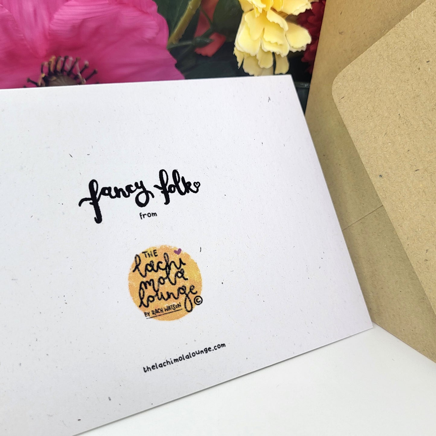 A white greeting card with black handwritten-style text reading 'fancy folk' and a circular gold logo for The Lachimolalounge, shown partly inside a kraft paper envelope with flowers in the background.