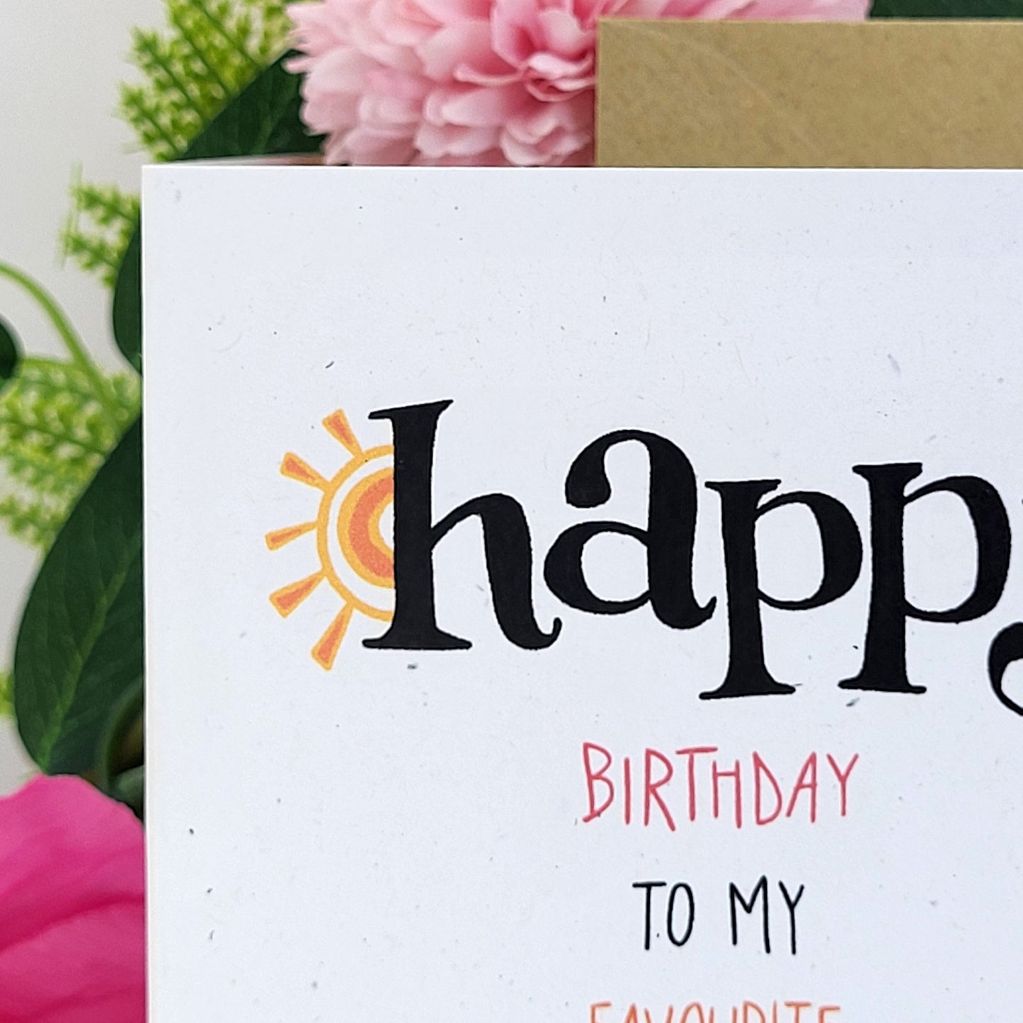 A close-up of a birthday card with the word 'happy' in large black script, a small orange and yellow sun graphic, and the text 'BIRTHDAY TO MY FAVOURITE' below. The card is styled with pink flowers and green leaves in the background.